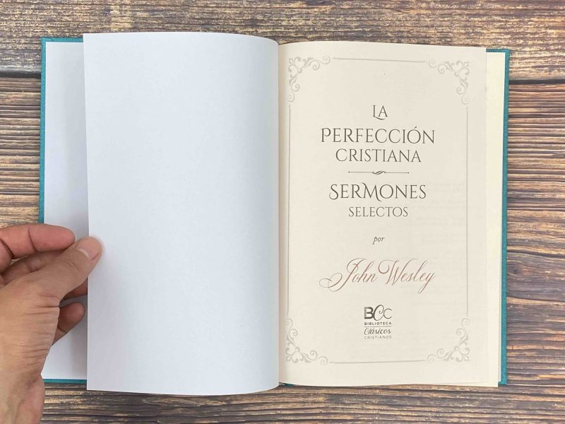 La imagen muestra una mano sosteniendo un libro abierto, con su cubierta exterior de color turquesa. La página de la derecha presenta el título La Perfección Cristiana Sermones Selectos y el nombre del autor John Wesley, junto con el logotipo de la Biblioteca Clásicos Cristianos en la parte inferior. La página izquierda está en blanco y el libro se encuentra sobre una superficie de madera que sugiere un ambiente tranquilo para la lectura y la reflexión.