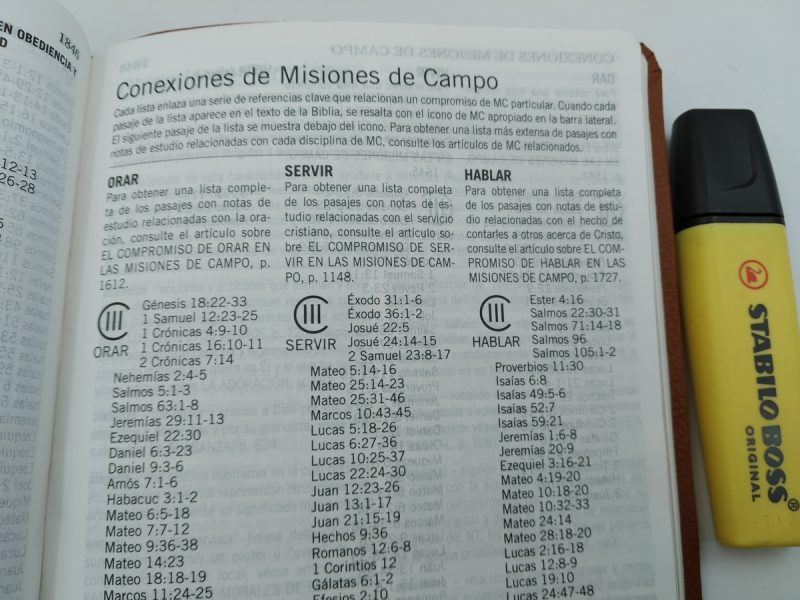 Fotografía de un libro abierto, posiblemente una Biblia, con una página titulada Conexiones de Misiones de Campo. El texto incluye referencias bíblicas organizadas bajo diferentes encabezados como ORAR, SERVIR y HABLAR. A la derecha del libro, descansa un marcador amarillo de la marca STABILO.