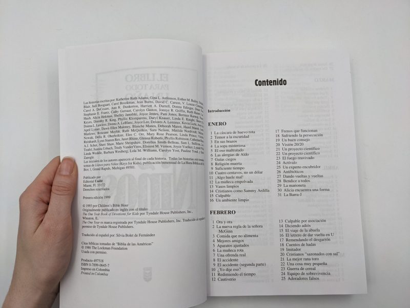 Una mano sosteniendo un libro abierto, mostrando una página con el título 'Contenido' y otra con detalles de publicación, en un fondo blanco.
