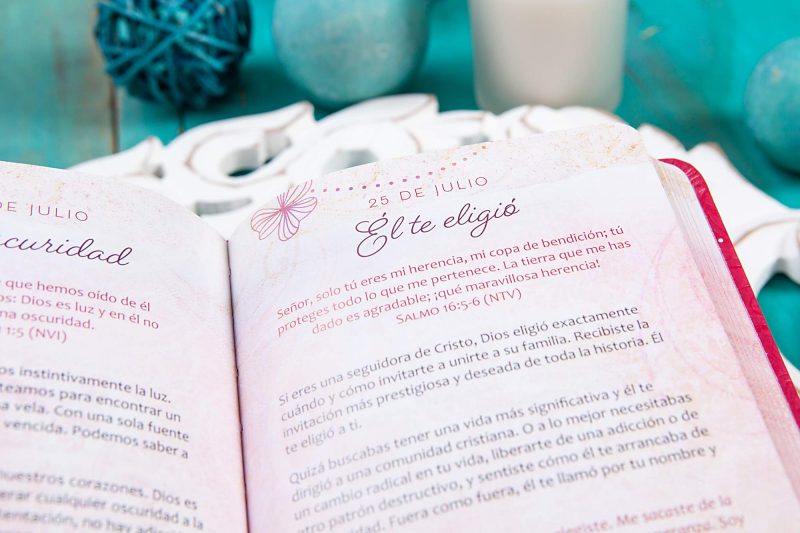 Un libro abierto mostrando una página con la fecha del 25 de julio y texto inspirador en español, rodeado de decoraciones de color turquesa y una vela.