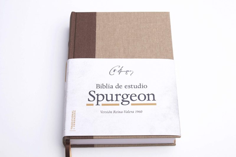 Una Biblia de estudio titulada Spurgeon con la versión Reina-Valera 1960, de tapa dura en colores blanco y marrón, reposando sobre un fondo blanco.