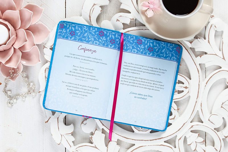 Una taza de café al lado de un libro abierto sobre una mesa de madera adornada con una flor de cerámica y un rosario de perlas.
