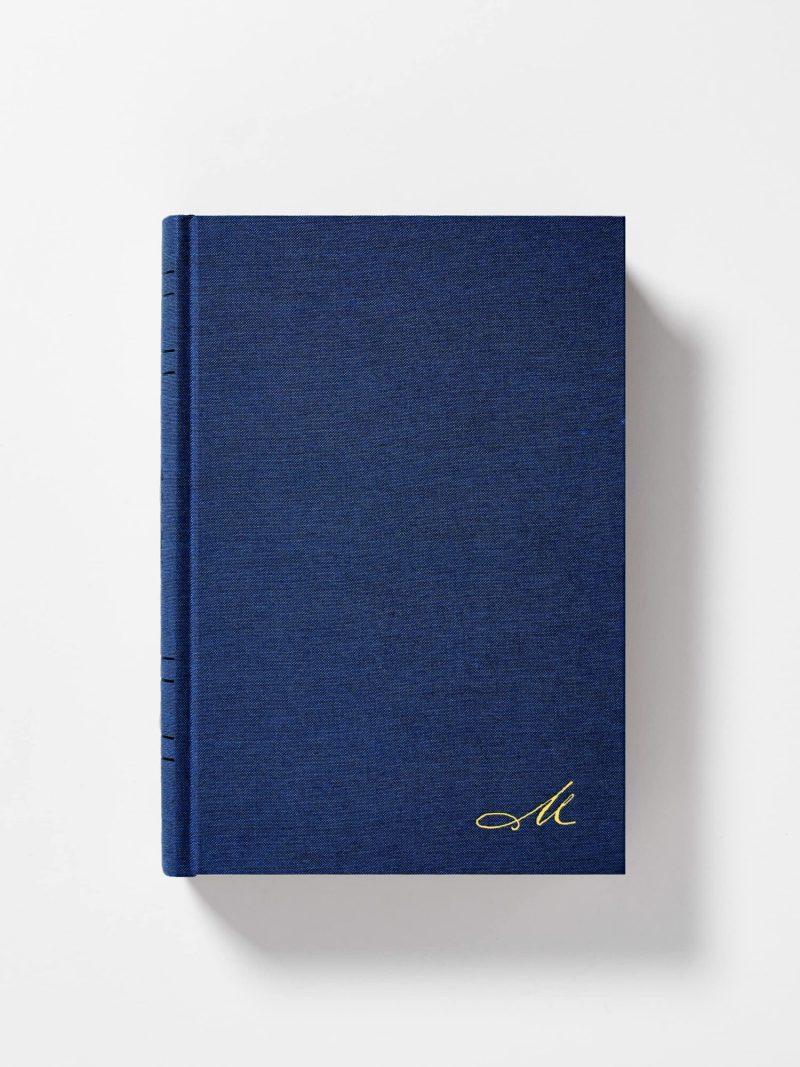 un libro de tapa dura con cubierta de tela texturizada en un tono azul profundo. En la esquina inferior derecha de la portada hay un monograma distintivo bordado en hilo dorado. El lomo del libro se distingue por líneas sutiles que sugieren una encuadernación clásica. El libro se presenta contra un fondo blanco neutro, lo que destaca su diseño simple pero sofisticado.