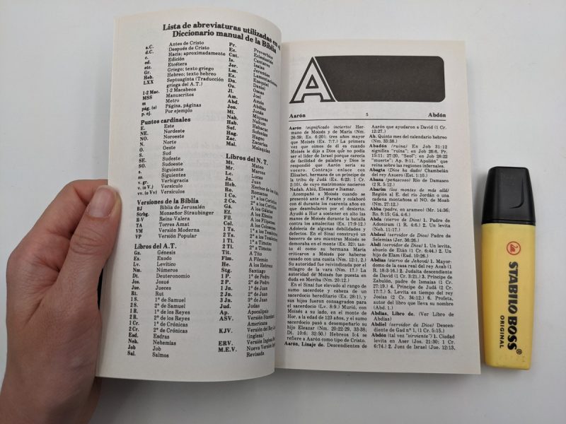 Una mano sostiene un diccionario bíblico abierto en la página de abreviaturas y el inicio de la sección 'A', junto a un marcador amarillo de texto Stabilo Boss.