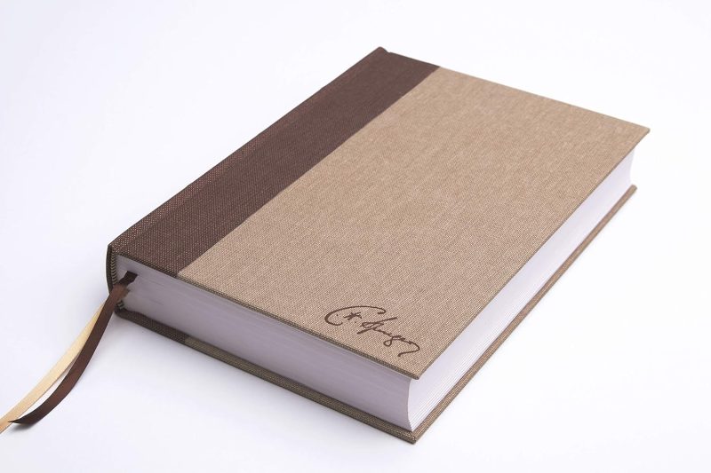 ** Un libro cerrado con tapa dura de dos tonos: marrón oscuro y beige, y un marcador de cinta marrón. La cubierta tiene una firma en la esquina inferior derecha.