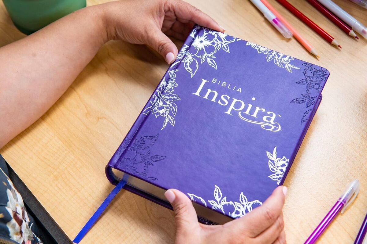 ** Persona sosteniendo una Biblia Inspira de tapa morada decorada con motivos florales sobre una mesa con lápices de colores.