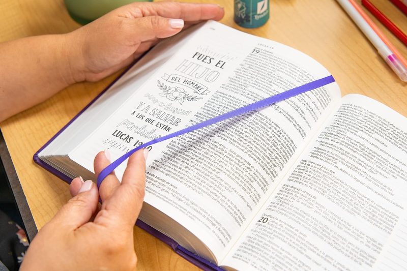 ** Manos sosteniendo un libro abierto con texto religioso y una cinta violeta como separador.