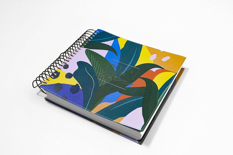 ** Cuaderno de espiral con una tapa de diseño abstracto y colorido, predominando tonos verdes, amarillos y azules sobre un fondo blanco.