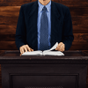 Un hombre de traje y corbata azules se encuentra detrás de un podio de madera sosteniendo un libro abierto contra un fondo de paneles de madera oscura.