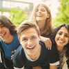 Cuatro amigos jóvenes sonriendo y divirtiéndose al aire libre, con dos de ellos cargando a los otros en sus espaldas en un entorno urbano verde.