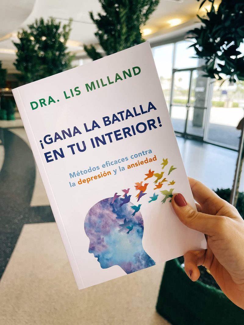 Una mano sujetando un libro titulado '¡GANA LA BATALLA EN TU INTERIOR!' por la Dra. Lis Milland. La portada muestra la silueta de un perfil humano con un fondo de acuarela y aves que parecen emerger volando desde la cabeza, simbolizando libertad o escape.