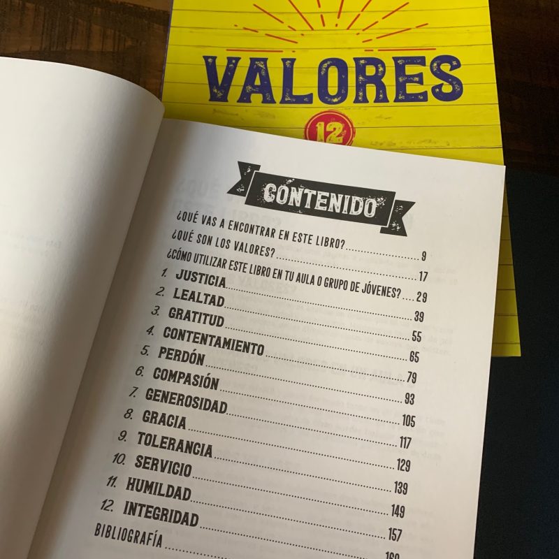 Página abierta de un libro mostrando el índice con el título 'CONTENIDO' y capítulos enumerados sobre diferentes valores como Justicia, Lealtad y Gratitud. El libro está abierto en una mesa de madera y la tapa superior se ve al fondo con el texto 'VALORES' y el número 12 en un círculo.