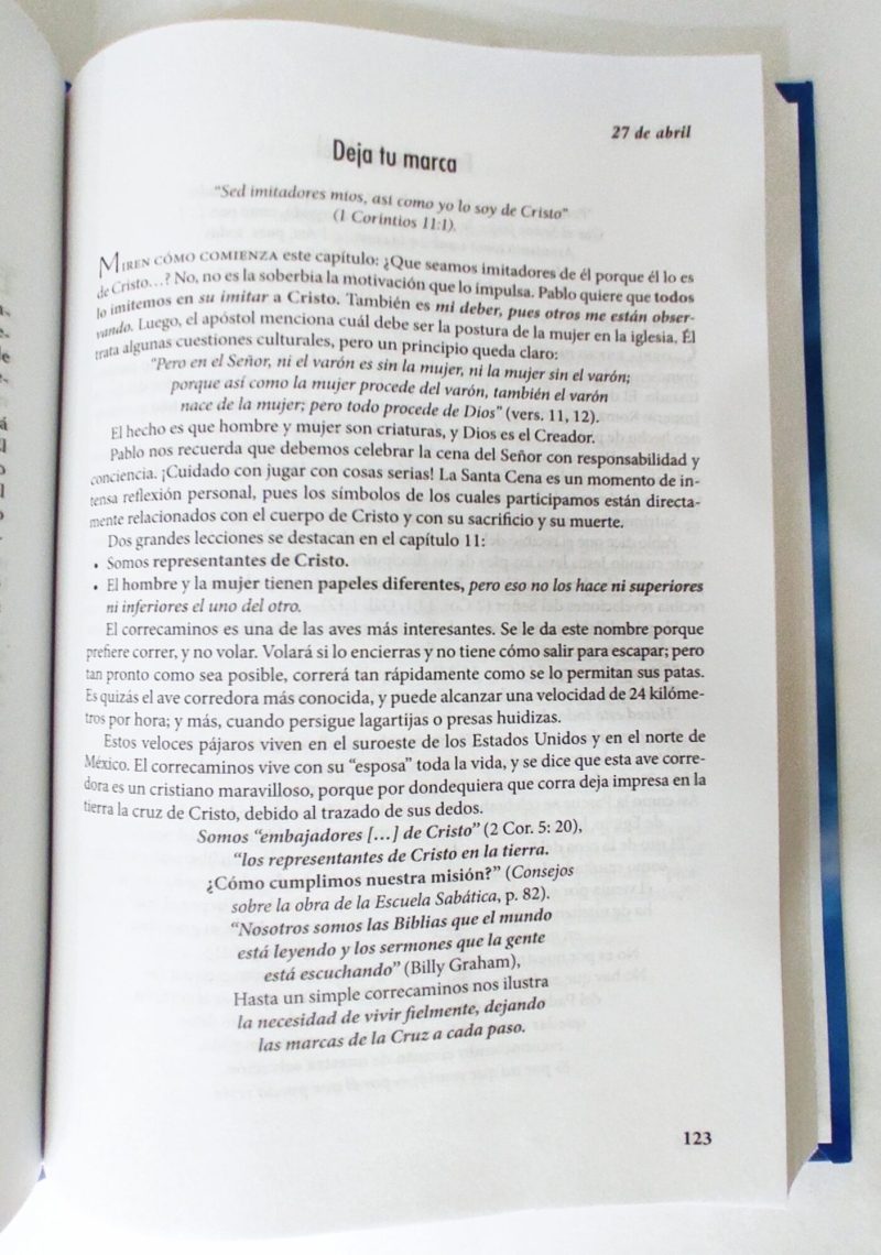 La imagen muestra un libro abierto en una página que incluye reflexiones espirituales o religiosas con un enfoque cristiano, dado que contiene citas bíblicas como 1 Corintios 111 y menciones de figuras como Billy Graham. El texto visible discute conceptos como la imitación de Cristo, los roles de hombres y mujeres en la perspectiva cristiana y la necesidad de dejar una marca positiva inspirada por la fe. Un marcador azul sobresale en el borde superior del libro, dando a entender que el lector ha marcado la página para continuar la lectura más tarde.