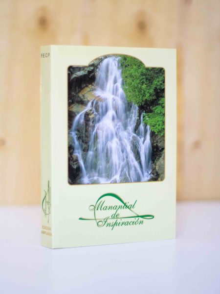 La imagen muestra un libro vertical con una portada que presenta una fotografía de una grandiosa cascada rodeada de frondosa vegetación, lo que sugiere un ambiente natural y sereno. El título del libro, Manantial de Inspiración, está escrito con una tipografía elegante en color verde oscuro que complementa el tema natural de la fotografía. En la esquina inferior izquierda de la cubierta, se puede ver la inscripción EDICION AMPLIADA en letras más pequeñas, indicando que esta versión del libro podría contener contenido adicional o actualizado. La imagen del libro está enfocada y se destaca contra un fondo de madera suave y difuso, que junto con la superficie blanca sobre la que descansa, crea un contraste que realza la presencia del libro.