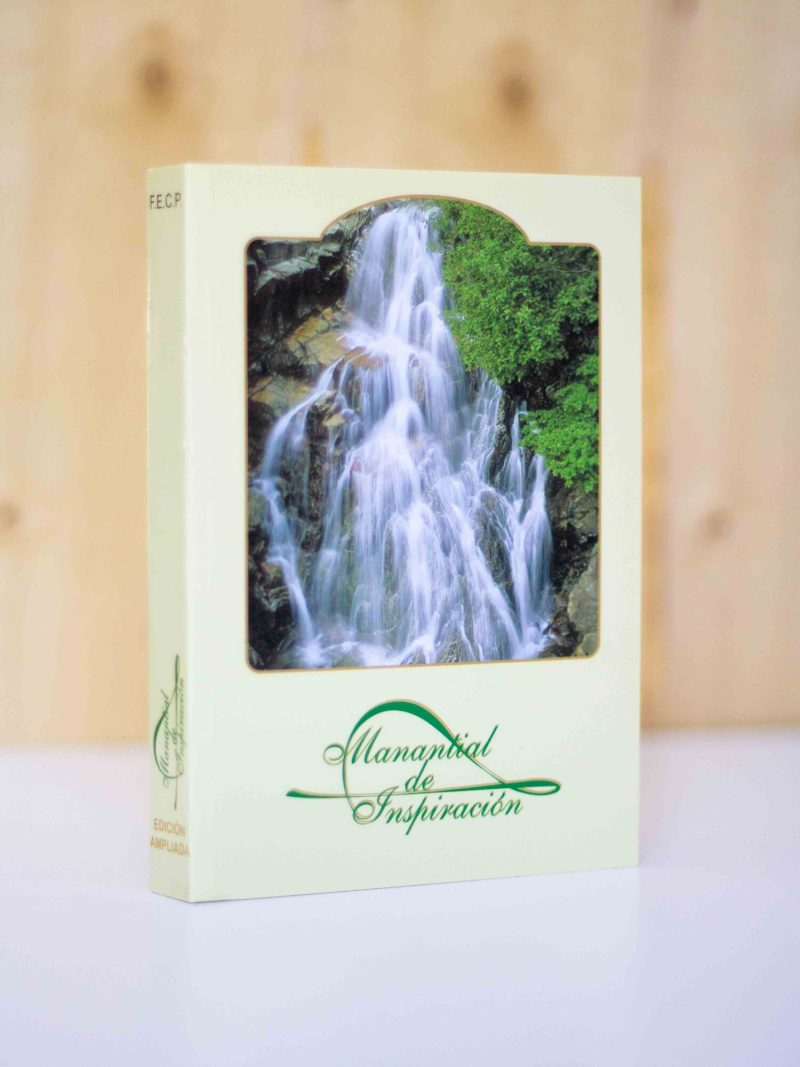 La imagen muestra un libro vertical con una portada que presenta una fotografía de una grandiosa cascada rodeada de frondosa vegetación, lo que sugiere un ambiente natural y sereno. El título del libro, Manantial de Inspiración, está escrito con una tipografía elegante en color verde oscuro que complementa el tema natural de la fotografía. En la esquina inferior izquierda de la cubierta, se puede ver la inscripción EDICION AMPLIADA en letras más pequeñas, indicando que esta versión del libro podría contener contenido adicional o actualizado. La imagen del libro está enfocada y se destaca contra un fondo de madera suave y difuso, que junto con la superficie blanca sobre la que descansa, crea un contraste que realza la presencia del libro.