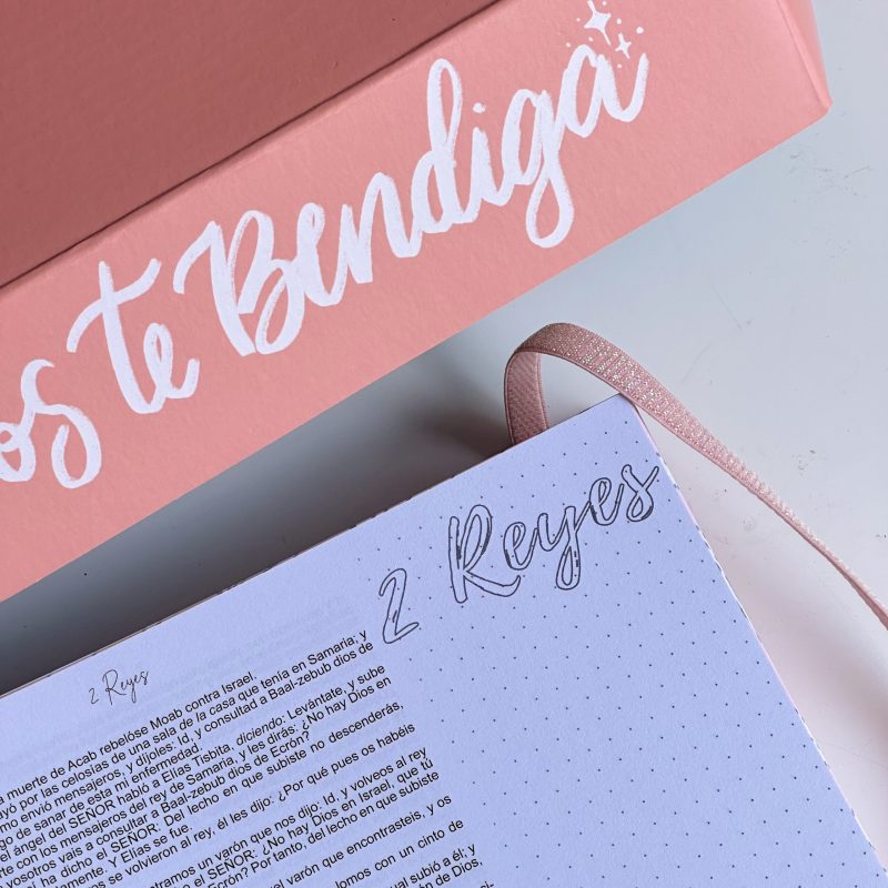 ** Una caja rosa con la frase "Dios te Bendiga" junto a un cuaderno abierto en la página titulada "2 Reyes".