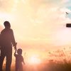 Silueta de un adulto y un niño de la mano ante una cruz grande, con el sol poniente y pájaros volando en el fondo.