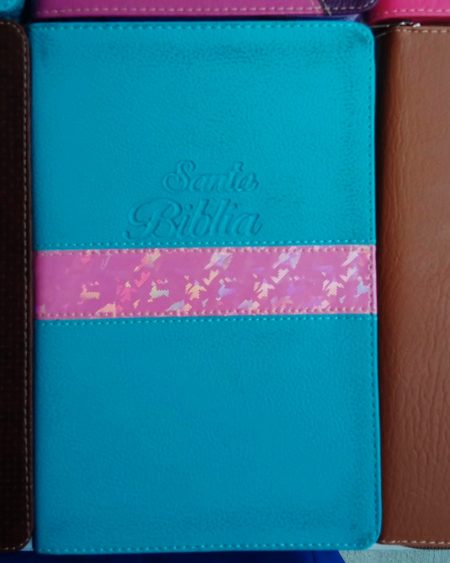 Vista cercana de una Biblia con cubierta turquesa y una banda decorativa rosa con cruces, junto a otras Biblias de diferentes colores.