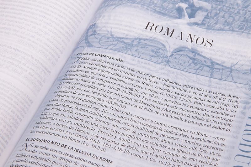 Página abierta de una Biblia mostrando el título de 'Romanos' con texto en español subrayado por una banda de diseño que contiene ilustraciones de columnas romanas.