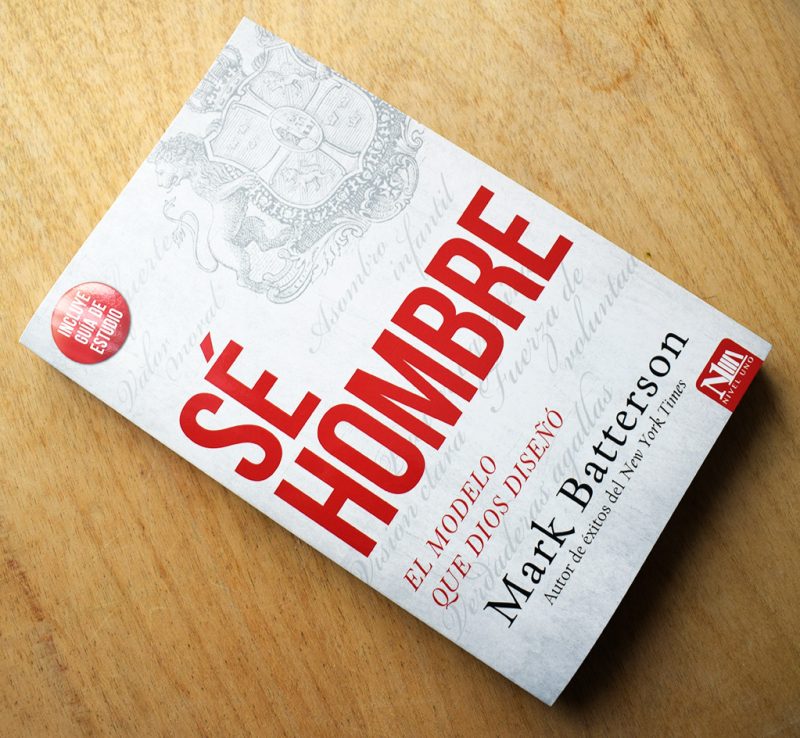 Portada del libro 'SÉ HOMBRE' de Mark Batterson, con un diseño en tonos rojos y grises, detallando el título y el autor, yaciente sobre una superficie de madera.