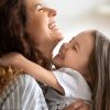 Una mujer sonriente abrazando cariñosamente a una niña pequeña, ambas riendo con alegría en un entorno doméstico.