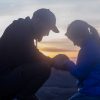 La imagen capta un momento sereno entre dos individuos sentados uno frente al otro mientras se toman de las manos, comunicando cercanía y conexión. El atardecer proporciona un ambiente tranquilo y pintoresco detrás de ellos, con el cielo irrumpiendo en una mezcla de colores cálidos que van desde el amarillo pálido hasta el azul oscuro. El paisaje que se vislumbra a la distancia sugiere una vista elevada y despejada, posiblemente una colina o una montaña, que añade un elemento de tranquilidad y aislamiento a la escena. La vestimenta de las personas sugiere que es un día fresco o de principios de otoño, y ambas llevan gorras y ropa abrigada. Su postura y el contacto físico transmiten afecto y un fuerte vínculo entre ellos.