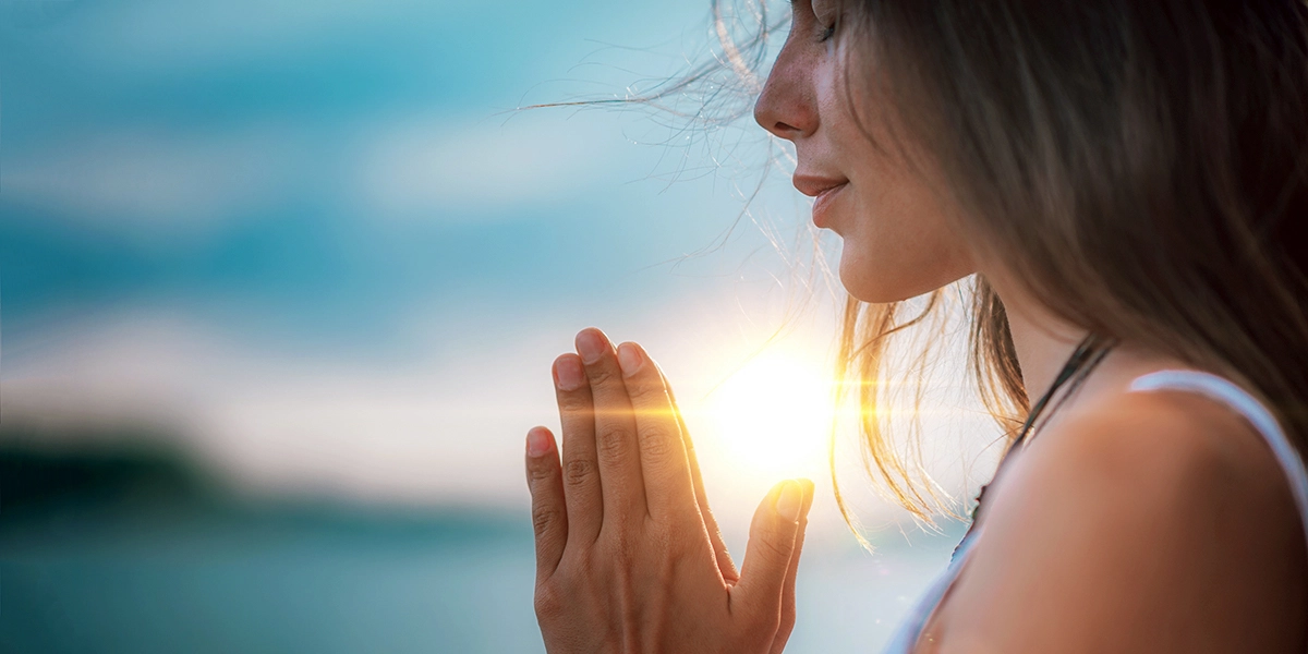 Una mujer en perfil capturada en un tranquilo momento de reflexión, con sus manos juntas y un suave atardecer que ilumina su rostro con destellos de luz solar.