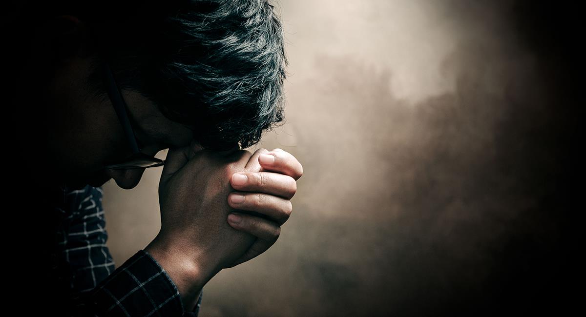 Una persona con gafas y una camisa a cuadros sostiene sus manos en gesto de reflexión o preocupación, con la cabeza inclinada y los ojos cerrados, contra un fondo oscuro y texturizado.