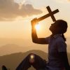 La imagen muestra una silueta de una persona sentada en el suelo, con el perfil hacia la cámara, sosteniendo una cruz de madera justo frente al sol, que se está poniendo en el horizonte. La luz del atardecer crea un efecto dramático, destacando la cruz y la persona contra un cielo anaranjado y amarillo, con capas sucesivas de montañas en la lejanía. El sol brilla intensamente y está parcialmente oculto detrás de la cruz, creando un halo de luz que resalta la escena de reflexión o plegaria. La persona parece estar en un momento de meditación o oración, tranquilamente inmersa en el entorno natural.