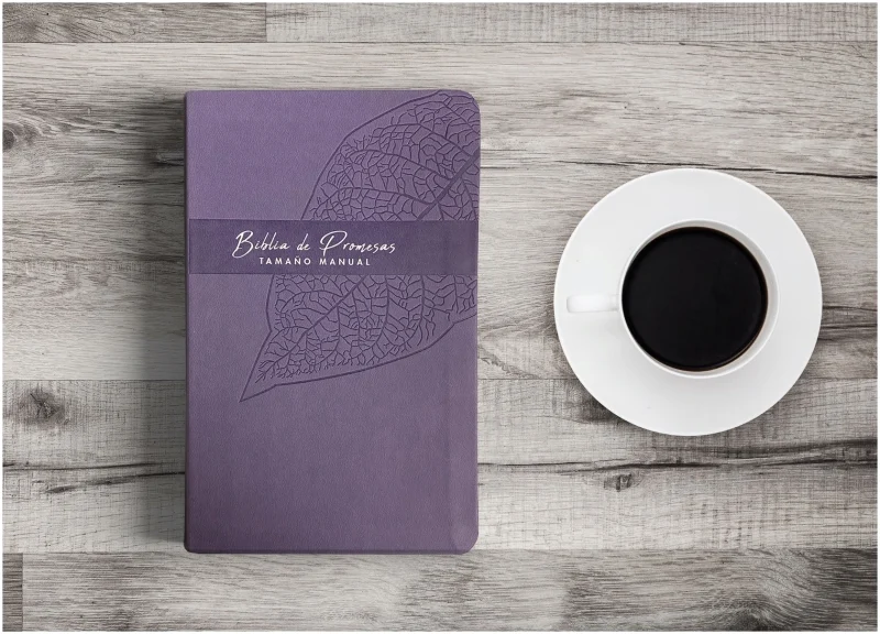 ** Un cuaderno de color morado oscuro con un diseño de hoja en la portada, colocado sobre una mesa de madera clara, acompañado de una taza de café negro.
