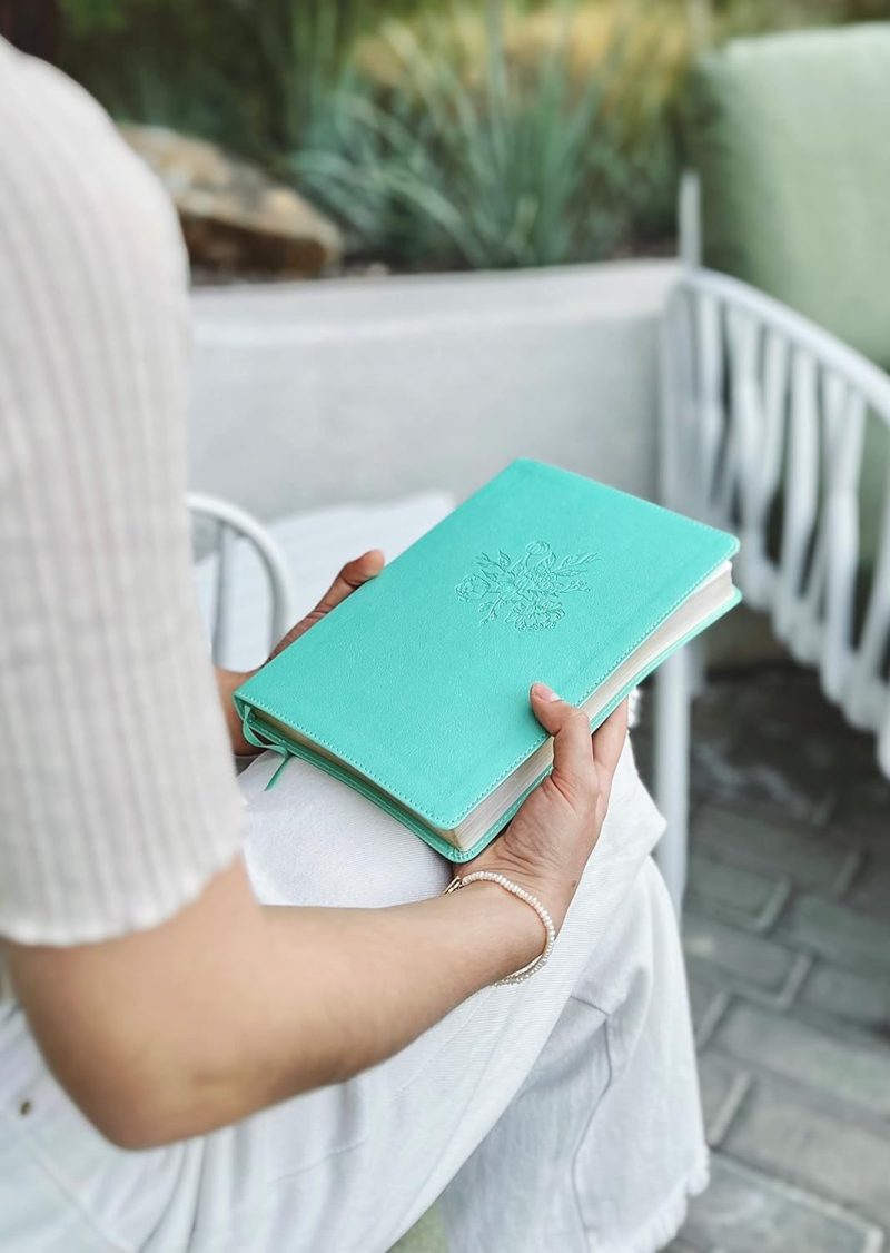 ** Persona sosteniendo un libro de portada verde turquesa mientras está sentada al aire libre en una silla.