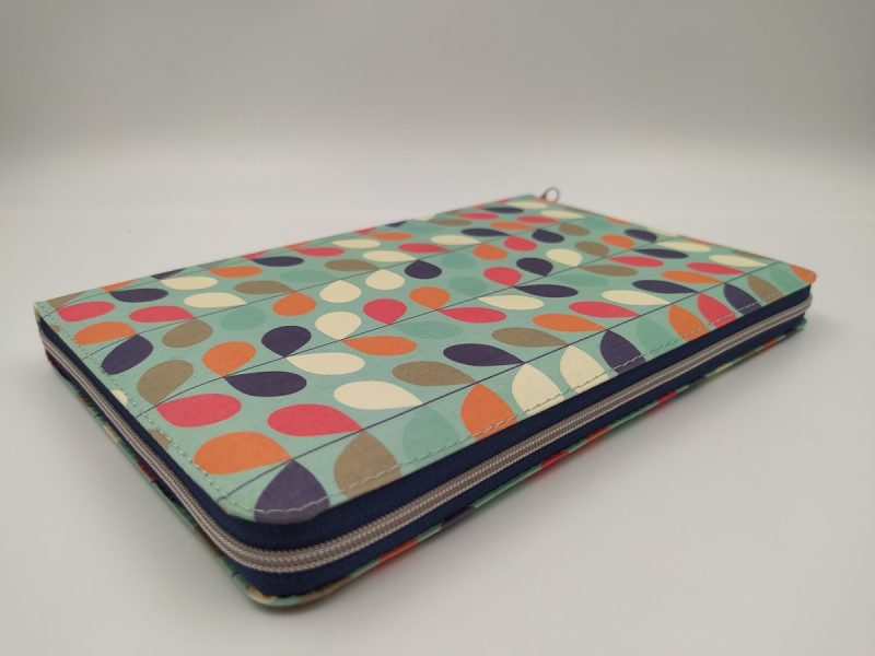 ** Un estuche plano con patrón de hojas coloridas en tonos verdes, azules, naranjas y cremas sobre un fondo verde claro, cerrado con una cremallera azul.