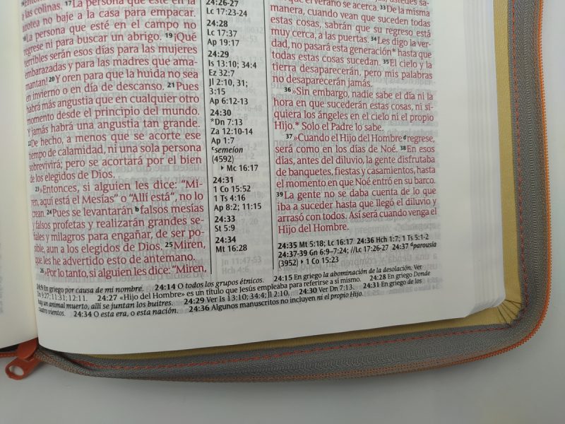 ** Primer plano de una página abierta de la Biblia con texto en rojo y negro. Observa columnas de escrituras y notas a pie de página.