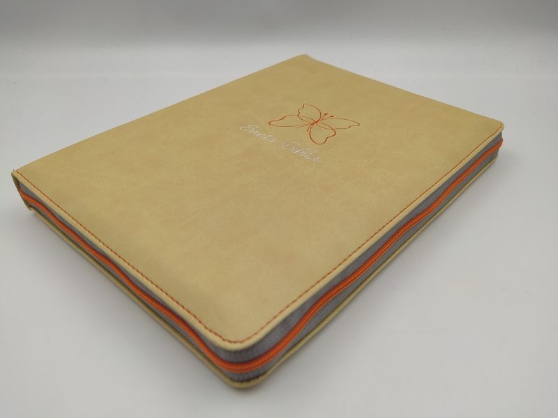 ** Cuaderno beige con un diseño de mariposa en la portada. Está cerrado y colocado sobre una superficie blanca.