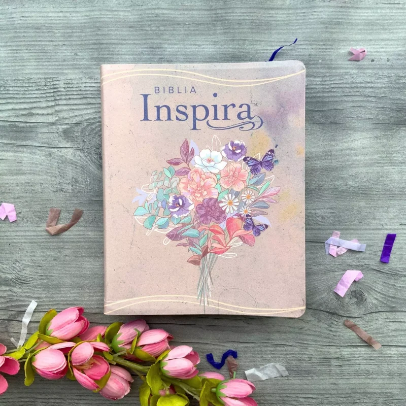 ** Biblia Inspira de tapa rosada con ilustración de un ramo de flores coloridas, sobre fondo de madera y decorada con flores rosas.