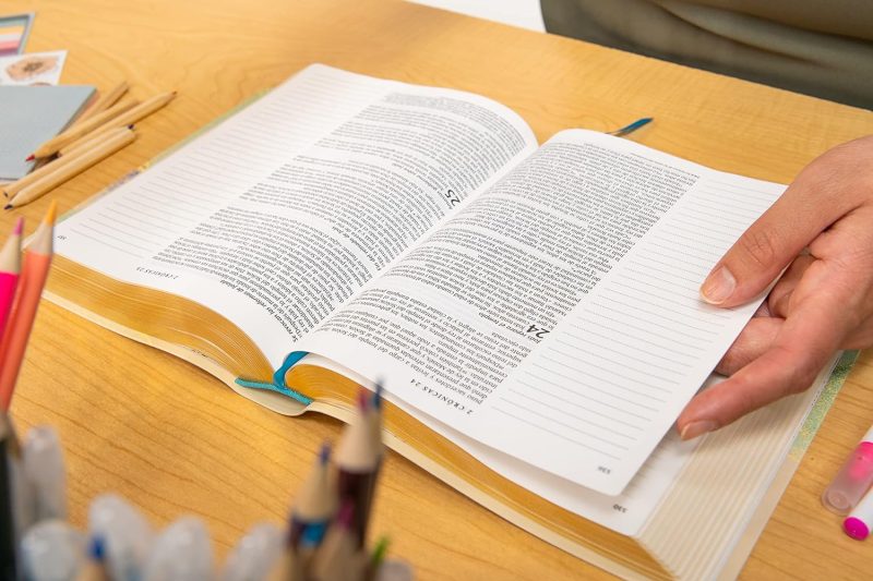 ** Persona hojeando un libro abierto sobre una mesa de madera, rodeado de lápices de colores.