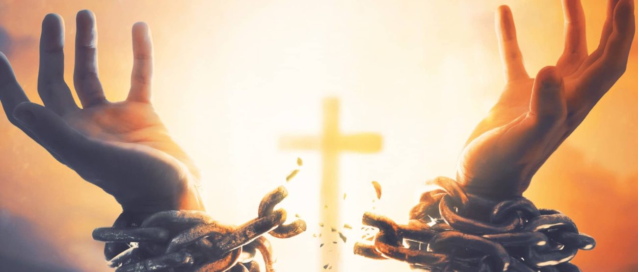 Manos liberadas de cadenas frente a la cruz de Cristo como símbolo de fortaleza y libertad.