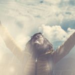 Una mujer con gafas y una chaqueta amarilla está de pie bajo un cielo nublado con rayos de luz atravesando las nubes, sus brazos levantados en una expresión de alegría o alabanza.