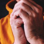 Primer plano de unas manos unidas en oración, visiblemente con nudillos y venas, sobre un fondo oscuro, sugiriendo concentración y súplica.