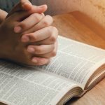 Manos entrelazadas en posición de oración o reflexión, apoyadas sobre una Biblia abierta con texto visible, iluminada por una luz lateral.
