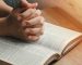 Manos entrelazadas en posición de oración o reflexión, apoyadas sobre una Biblia abierta con texto visible, iluminada por una luz lateral.