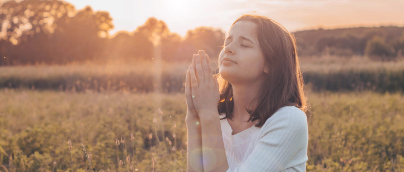 El perdón sin límites: ¿Qué significa “70 veces 7” en la Biblia? 4 Mujer de pie en un campo al atardecer con las manos juntas en oración
